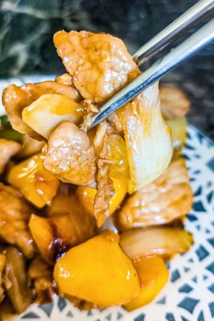 metal chopsticks holding pork and onion from a helping of sweet and sour pork on a blue and white plate