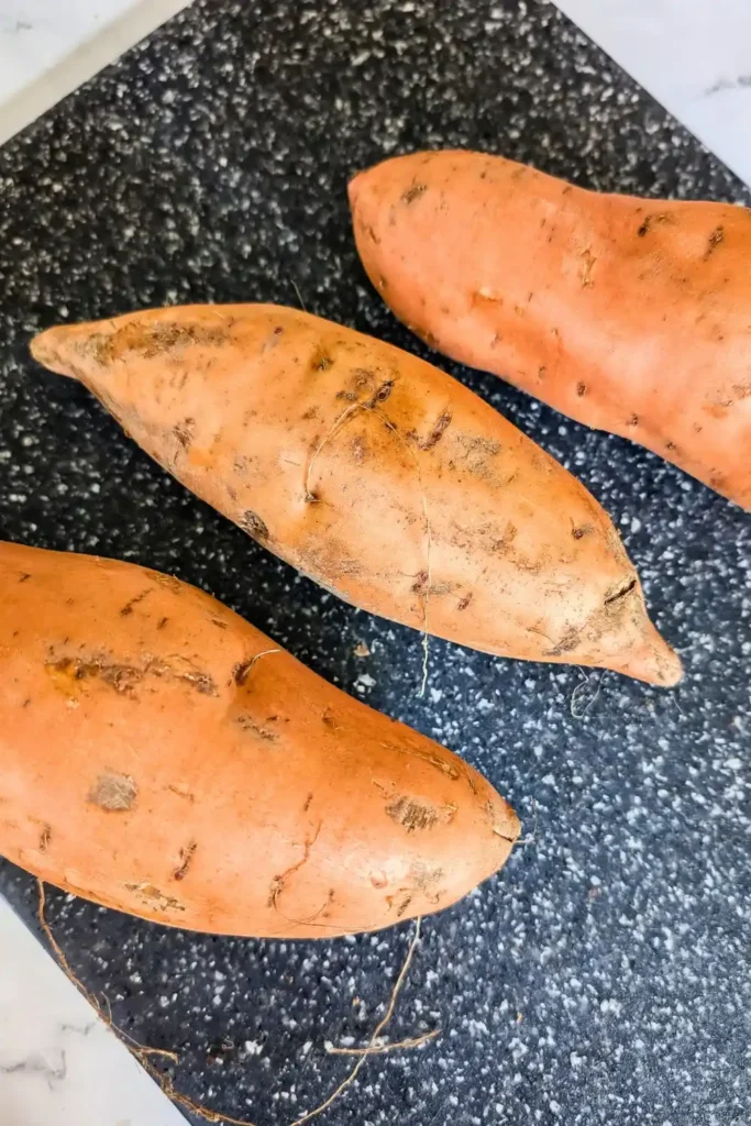 whole sweet potatoes sit on black speckled cutting board