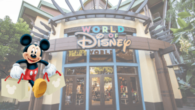 An entrance to the World of Disney store with a large sign above the doors. A joyful Mickey Mouse, holding shopping bags, is superimposed on the left side of the image, welcoming visitors. The store features colorful architectural elements and various Disney merchandise inside.