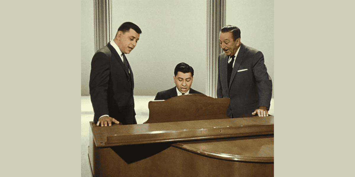 Three men are seen around a piano. One man is seated at the piano, playing, while the other two stand on either side of him, appearing engaged and possibly singing. The background is plain with vertical lines, suggesting a studio setting where they might be inspired by Walt Disney quotes.