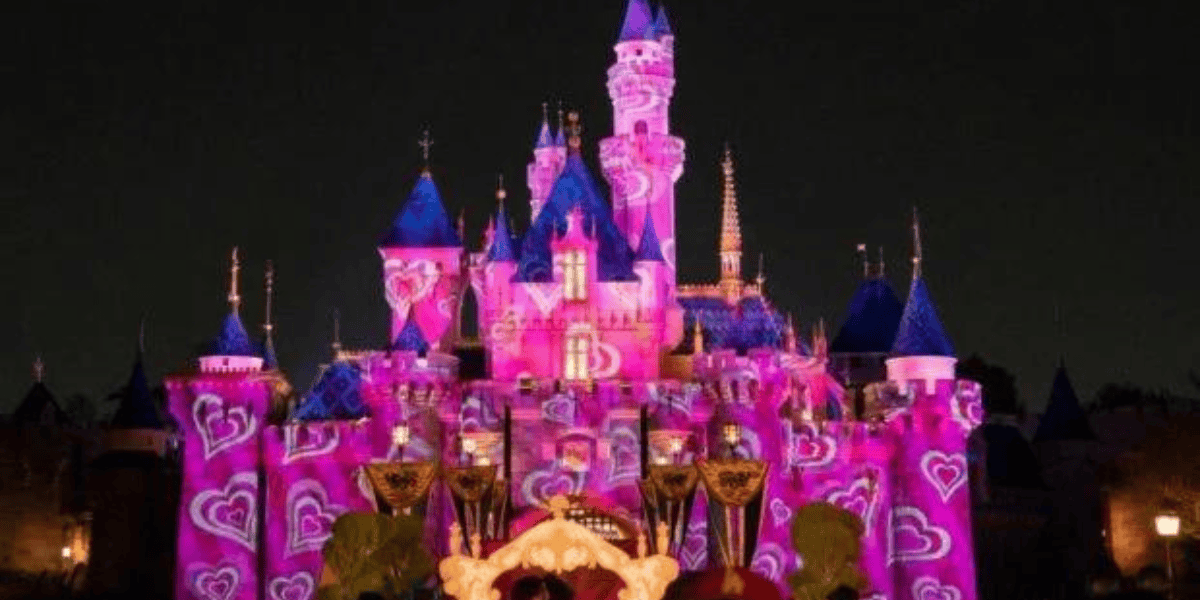 A grand castle illuminated at night, featuring vibrant pink and purple lights with heart patterns projected onto the structure. The towers are topped with blue spires, and the scene is set against a dark sky, creating a magical, enchanting atmosphere.