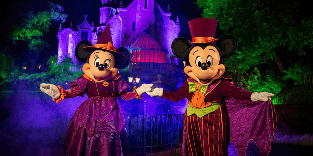Mickey and Minnie Mouse are dressed in Halloween-themed costumes—Minnie in a purple witch outfit and hat, Mickey in a purple suit with an orange vest and green bow tie. They are holding hands and posing in front of a spooky, illuminated castle surrounded by mist.