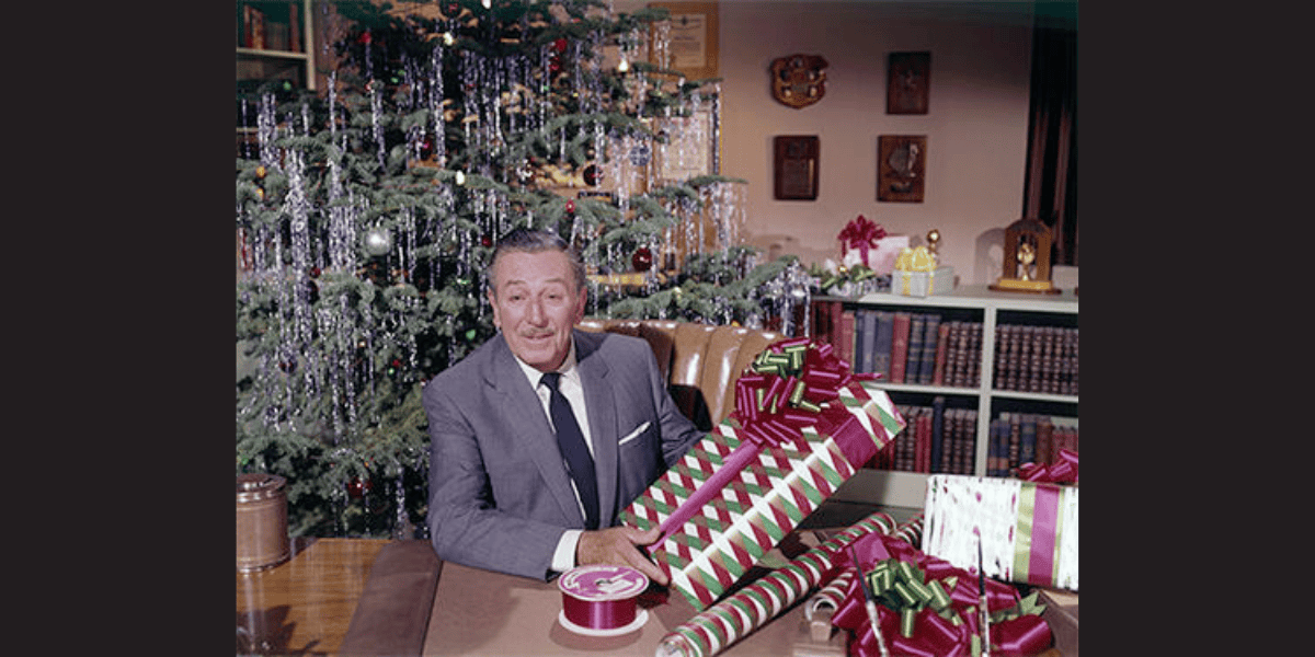 A man in a suit sits at a table wrapping Christmas gifts, embodying the magic often captured by Walt Disney quotes. Behind him, a decorated Christmas tree with tinsel and ornaments adds to the festive spirit. The table is filled with wrapped presents and ribbons in vibrant colors, with shelves of books and decorative items in the background.