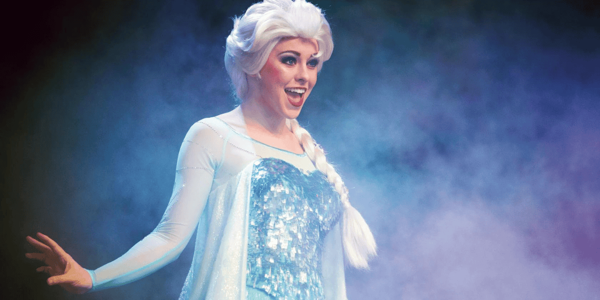 A person dressed as an ice queen character stands amidst a foggy, mystical background. They have platinum blonde hair styled in a braid, and are wearing a sparkly, light blue dress with sheer sleeves, smiling and extending one arm to the side.