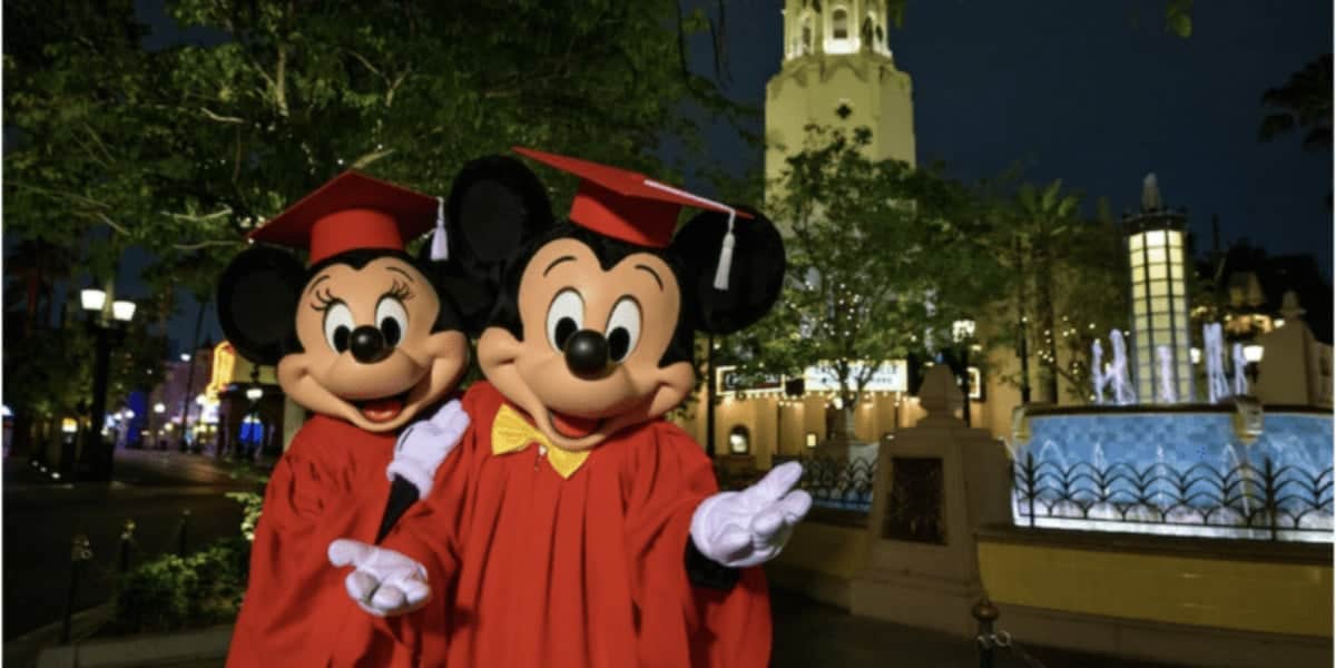 Mickey and Minnie Mouse in red graduation gowns and caps pose happily in front of a fountain and a tall building, surrounded by trees and lights, during the night.