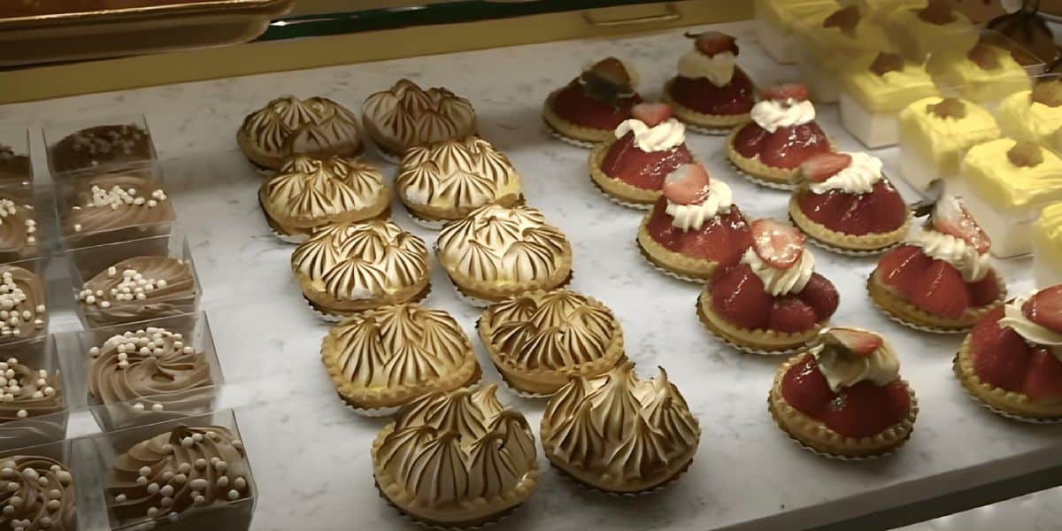 A bakery display showing an assortment of desserts: lemon meringue tarts and strawberry cream tarts with fresh strawberries, alongside chocolate cupcakes topped with creamy swirls and white sprinkles.