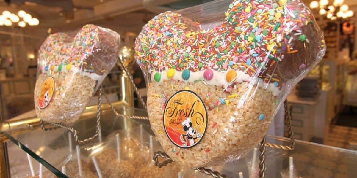 Two large, Mickey Mouse-shaped Rice Krispie treats are displayed on stands, covered with colorful sprinkles and candies. Each treat is wrapped in clear plastic and has a decorative label featuring a cartoon character.
