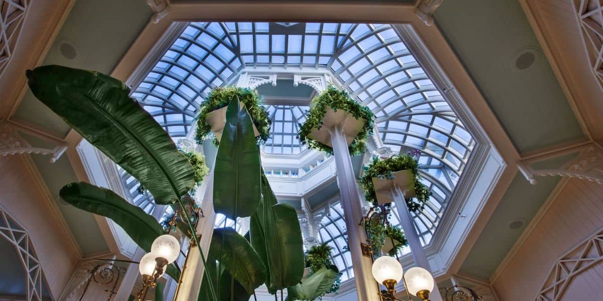 Looking up at a glass atrium ceiling with large tropical leaves and tall white columns adorned with lush green plants. Elegant lamps with round bulbs are visible in the foreground, enhancing the spacious and airy atmosphere.