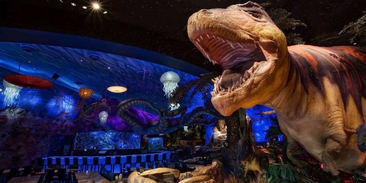 A vibrant themed restaurant interior featuring a large T-Rex model with its mouth open. The atmosphere is enhanced by dim lighting, glowing jellyfish decorations on the ceiling, and a long bar with stools in the background.