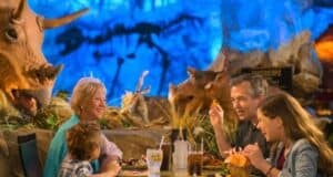 A family of four enjoys a meal at a themed restaurant with dinosaur decor. Two adult figures and two children are seated around a table, smiling and interacting. A triceratops statue is visible in the background against a blue-lit setting.