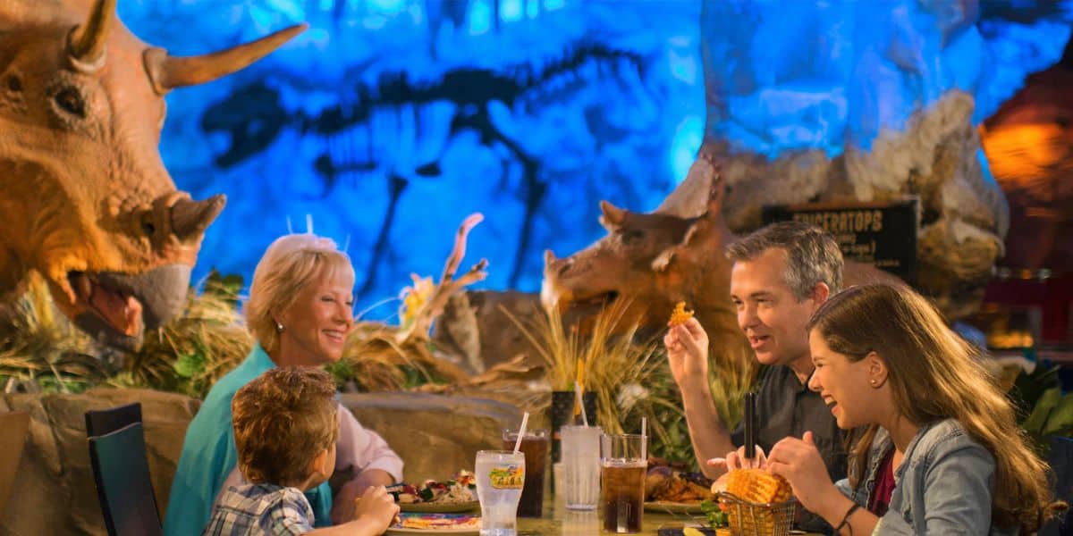 A family of four enjoys a meal at a themed restaurant with dinosaur decor. Two adult figures and two children are seated around a table, smiling and interacting. A triceratops statue is visible in the background against a blue-lit setting.
