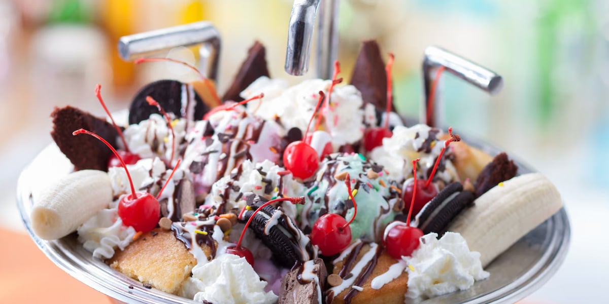 A large Disney World ice cream sundae in a sink, topped with whipped cream, cherries, chocolate sauce, and sprinkles, surrounded by banana slices, brownies, and cookies.