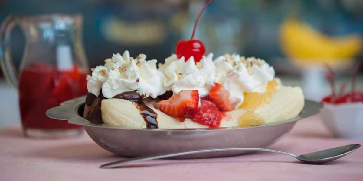 A banana split is elegantly served in a silver dish, featuring scoops of vanilla ice cream, whipped cream, a cherry, chocolate sauce, and sliced strawberries. There's a spoon beside the dish and a jug with red sauce in the background—reminiscent of Disney World ice cream treats.