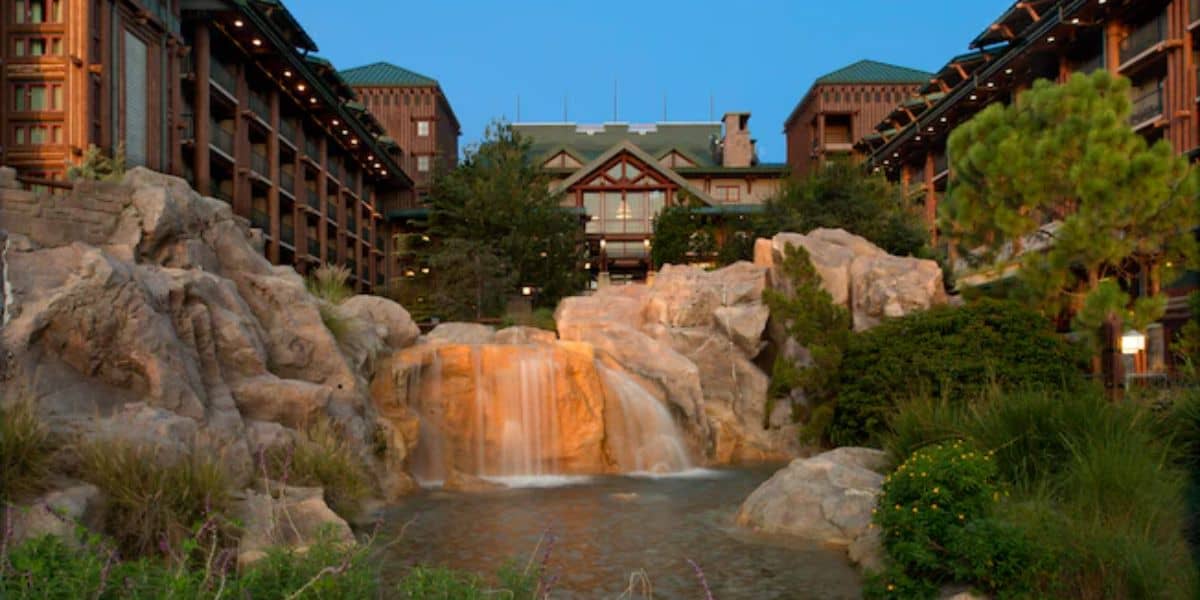 A rustic lodge with a triangular roof stands in the background, surrounded by lush greenery. In the foreground, a waterfall cascades over large rocks into a serene pool, creating a picturesque scene.