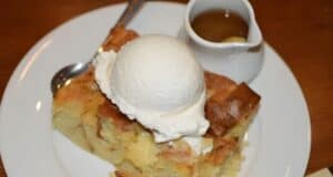 A slice of bread pudding topped with a scoop of vanilla ice cream is served on a white plate. A small jug of sauce and a spoon accompany the dessert.