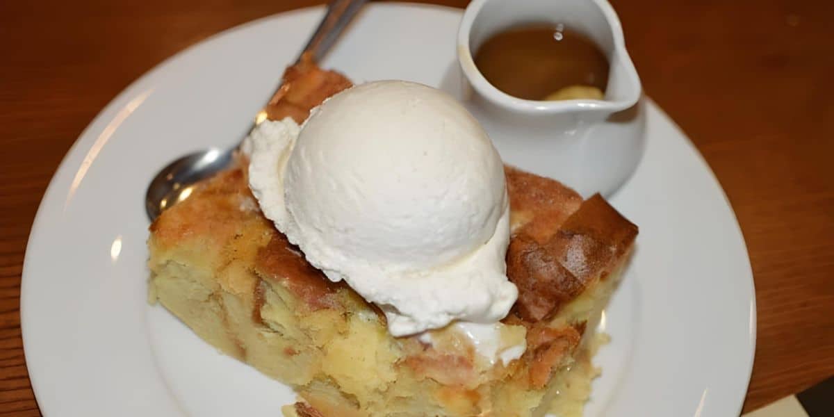 A slice of bread pudding topped with a scoop of vanilla ice cream is served on a white plate. A small jug of sauce and a spoon accompany the dessert.