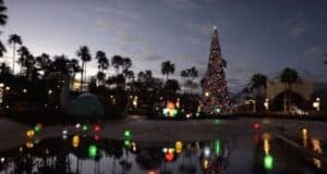 A festive, towering Christmas tree adorned with colorful lights stands amidst a park at dusk. Palm trees and glowing decorations are visible in the background, with vibrant lights reflecting on the water's surface in the foreground.