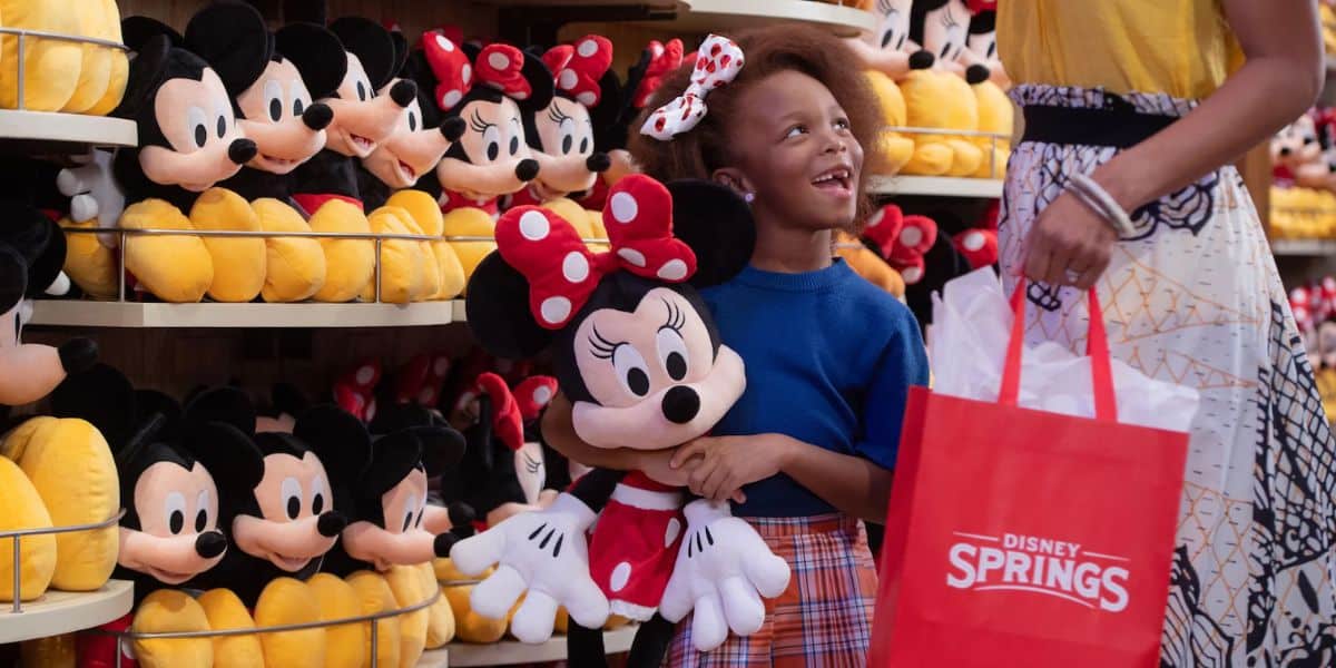 A girl happily holds a Minnie Mouse plush toy in a store filled with similar toys. She looks up at a person holding a red Disney Springs shopping bag. The shelves are packed with plush toys in the background.