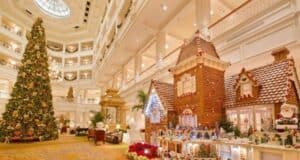 A grand hotel lobby beautifully decorated for Christmas. A large, ornate Christmas tree stands beside an elaborate gingerbread house adorned with festive decorations, including Santa Claus figures and holiday lights. The space is warm and inviting.