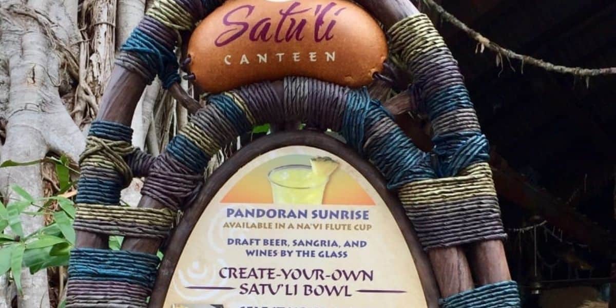 Sign for Satu'li Canteen featuring a menu with options for "Pandoran Sunrise," draft beer, sangria, wine, and "Create-Your-Own Satu'li Bowl." The sign is woven with colorful fabric and displayed outdoors among trees.