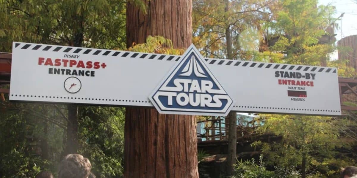 Sign for "Star Tours" at an amusement park entrance, featuring a "Disney FastPass+ Entrance" on the left with a clock, and "Stand-By Entrance" space on the right for wait time, surrounded by trees.
