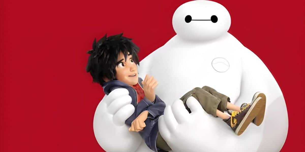 A large, white, inflatable robot gently holds a young boy with dark hair wearing a blue jacket and brown pants. They are against a bright red background. The robot’s expression is simple, with two dots for eyes and a line for a mouth.
