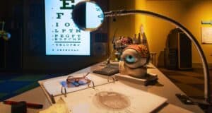 A whimsical desk scene with a large eye sculpture wearing glasses, a magnifying glass, an eye chart, and scientific papers. The room is dimly lit with a cozy, eclectic atmosphere.