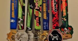 A collection of colorful race medals hanging in a row. Each medal features unique designs and event names, including marathons, a 10-miler, and holiday-themed races. Disney characters are pictured on some of the ribbons.