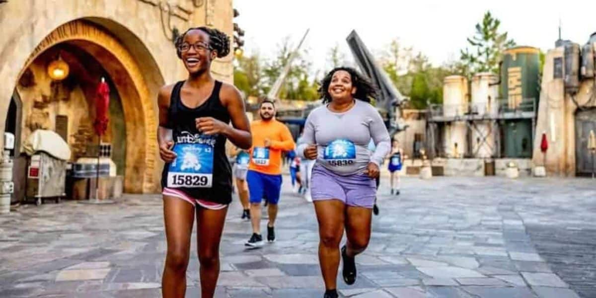 People participating in a themed fun run, smiling as they jog through a park area with rustic, fantasy-like structures in the background. They are wearing casual athletic clothes and race bibs.