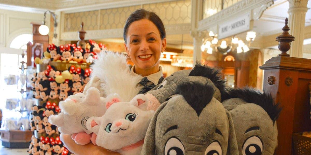 A person smiling while holding several plush toys, including characters resembling a white cat and a gray donkey. The setting appears to be a store with more plush toys displayed in the background.