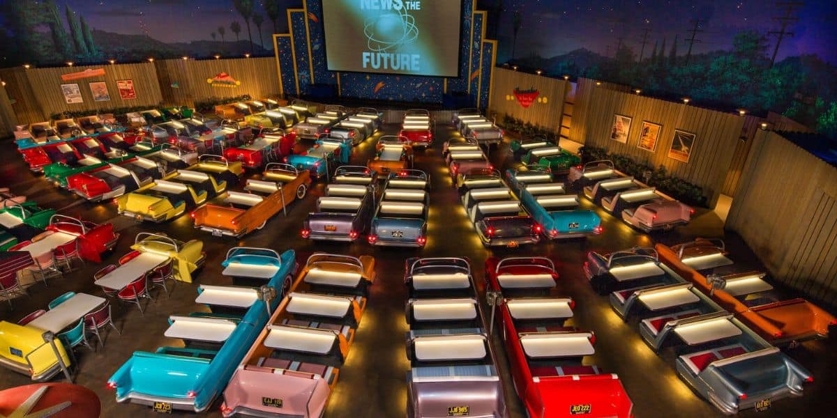 A retro-themed indoor cinema with colorful vintage car seats arranged in rows facing a large screen displaying "New is the Future." The cinema features a starry night sky backdrop, evoking an outdoor drive-in theater experience.
