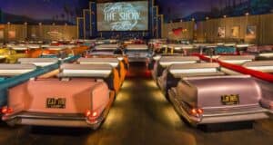 Rows of vintage car-shaped booths inside a retro-themed drive-in theater. A large screen displays "Enjoy the Show, Folks!" The atmosphere is cozy with dim lighting and starry decorations on the ceiling.