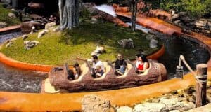 A group of people enjoy a ride in a log-shaped boat on a water theme park attraction. The ride winds through a green, rocky landscape with splashes of water creating an exciting atmosphere.