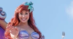 A woman dressed as a mermaid waves happily. She has long red hair, a shell-like top, and a hair accessory with seashells. She holds a fork in one hand against a bright blue sky.