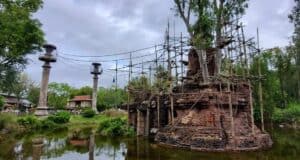 An ancient brick structure entwined with tree branches stands in a pond, surrounded by scaffolding and lush greenery. Tall columns with circular tops are visible nearby under a cloudy sky.