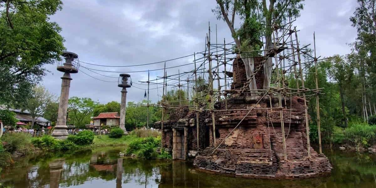 An ancient brick structure entwined with tree branches stands in a pond, surrounded by scaffolding and lush greenery. Tall columns with circular tops are visible nearby under a cloudy sky.