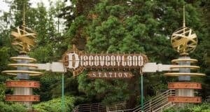 A retro-futuristic station entrance sign reading "Discoveryland Station" is flanked by two metallic, spherical sculptures with rings. The scene is set against a backdrop of green trees and surrounding foliage.