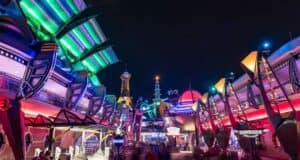 A vibrant, futuristic-themed amusement park area at night, featuring brightly lit buildings with neon lights in green, blue, and red. Crowds of people are blurred in motion, highlighting a lively atmosphere.
