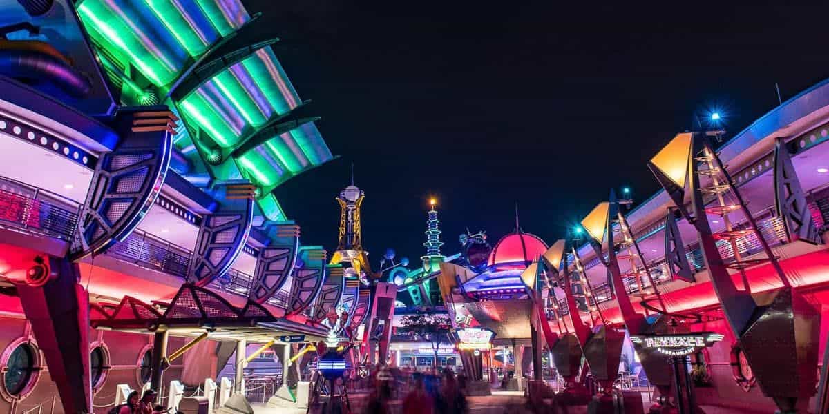 A vibrant, futuristic-themed amusement park area at night, featuring brightly lit buildings with neon lights in green, blue, and red. Crowds of people are blurred in motion, highlighting a lively atmosphere.