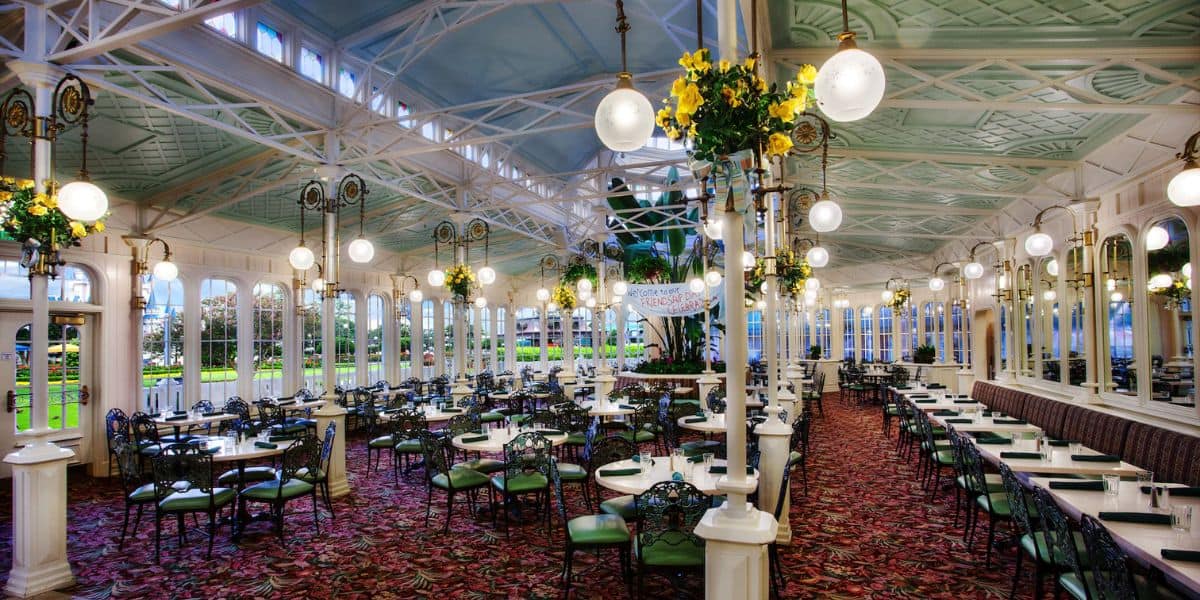 A spacious, elegant dining area with large windows, ornate light fixtures, and green-accented chairs. The ceiling is high with white and blue details, and there are floral arrangements hanging from pillars.