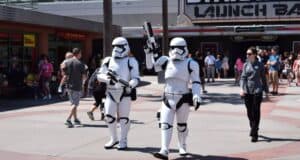 Two people in Stormtrooper costumes walk in front of a "Star Wars Launch Bay" sign at a theme park. They carry blasters as visitors in casual clothing stroll nearby. The scene is sunny and busy.