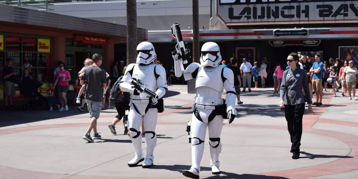 Two people in Stormtrooper costumes walk in front of a "Star Wars Launch Bay" sign at a theme park. They carry blasters as visitors in casual clothing stroll nearby. The scene is sunny and busy.