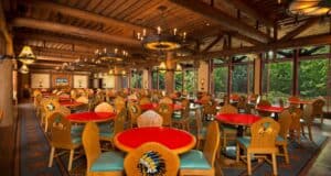 A rustic restaurant interior featuring wooden ceiling beams and Native American-themed decorations. Colorful, round tables with red tops are surrounded by wooden chairs decorated with feather motifs. Large windows reveal greenery outside.