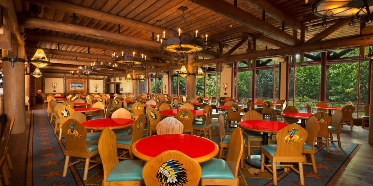 A rustic restaurant interior featuring wooden ceiling beams and Native American-themed decorations. Colorful, round tables with red tops are surrounded by wooden chairs decorated with feather motifs. Large windows reveal greenery outside.