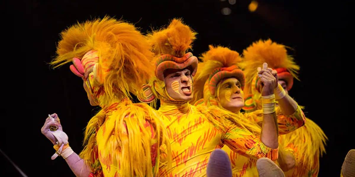 Four performers in vibrant lion costumes with orange and yellow fur and face paint, energetically pose on stage. The background is dark, highlighting their expressive, theatrical gestures.
