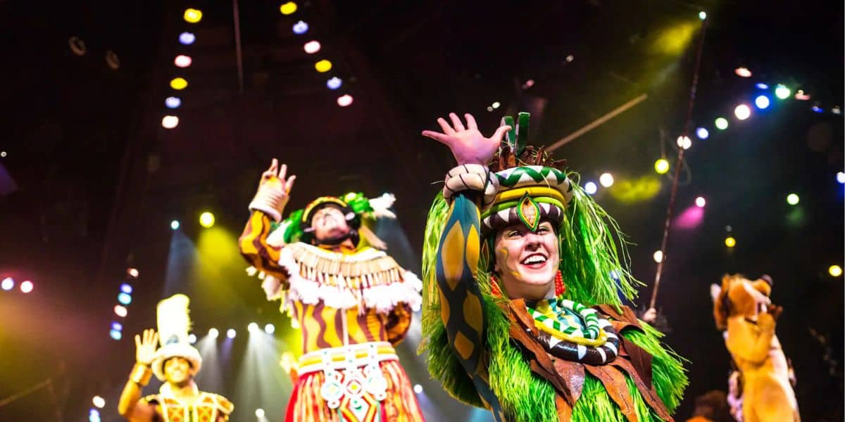 Performers in colorful costumes, including feathers and vibrant patterns, are on stage under bright, multicolored lights. They are smiling and waving their hands, creating an energetic and joyful atmosphere.