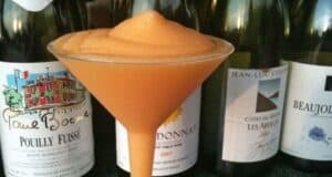 A slushy cocktail served in a martini glass, standing in front of a row of assorted wine bottles with colorful labels. The drink is orange and has a frothy texture.