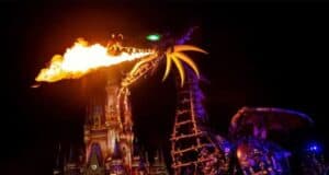 A large, mechanical dragon breathes fire in front of an illuminated castle during a nighttime parade. The dragon has glowing green eyes and a spiked metallic body, with a dark sky in the background.