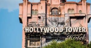 The image shows the Hollywood Tower Hotel, a tall, intricately designed building with a weathered facade and an ornate sign. The structure has a pinkish hue, and the sky is blue with a few clouds in the background. Trees partially obscure the view.