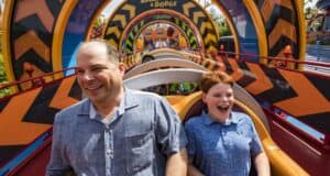 A man and a boy excitedly ride a vibrant roller coaster. The track features bold, colorful patterns creating a dynamic visual backdrop. Both are smiling widely, capturing the thrill and joy of the moment.
